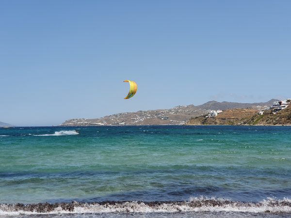 A kitesurfer out on the turquoise water off a Mykonos beach, with the town visible across the bay