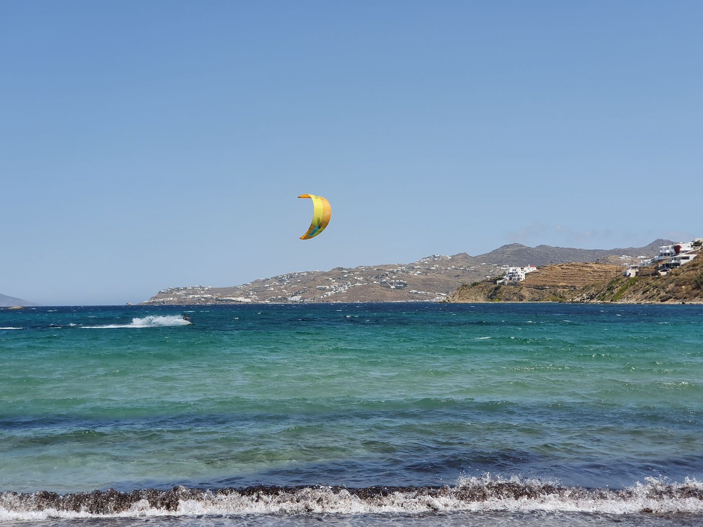 A kitesurfer out on the turquoise water off a Mykonos beach, with the town visible across the bay