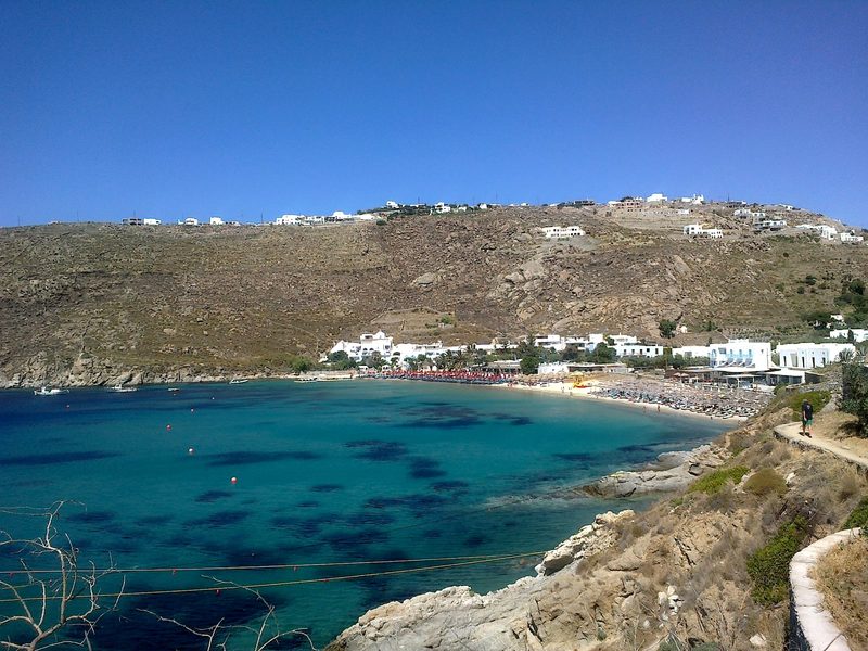 Sunbathers relax on Psarou Beach in Mykonos, with clear blue waters and white sand.