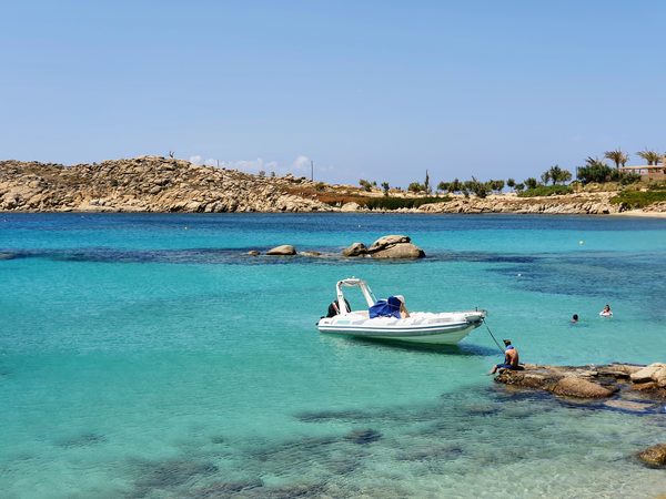 A small boat anchored in a turquoise cove below a rocky headland on the south coast of Mykonos