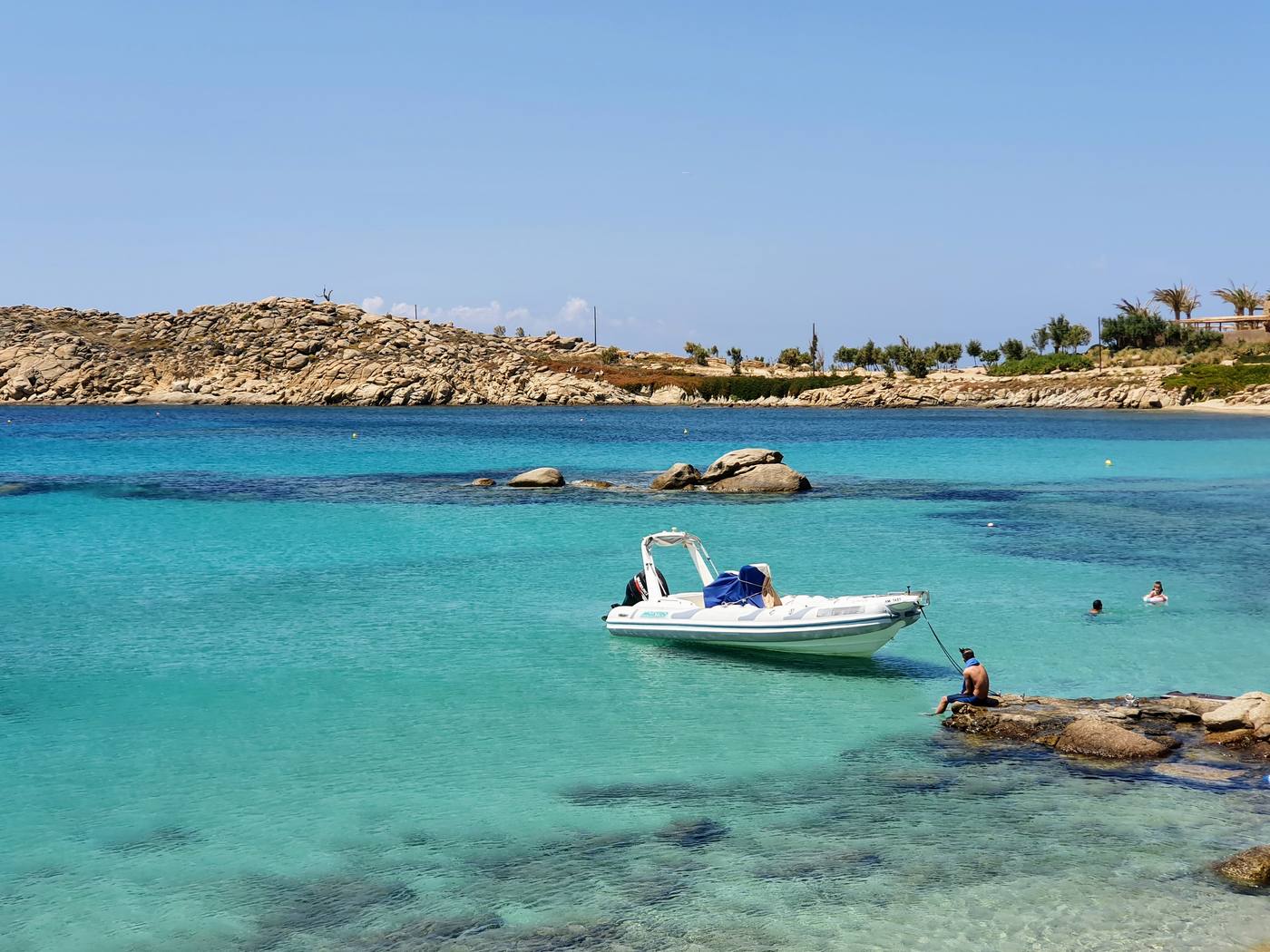 A small boat anchored in a turquoise cove below a rocky headland on the south coast of Mykonos