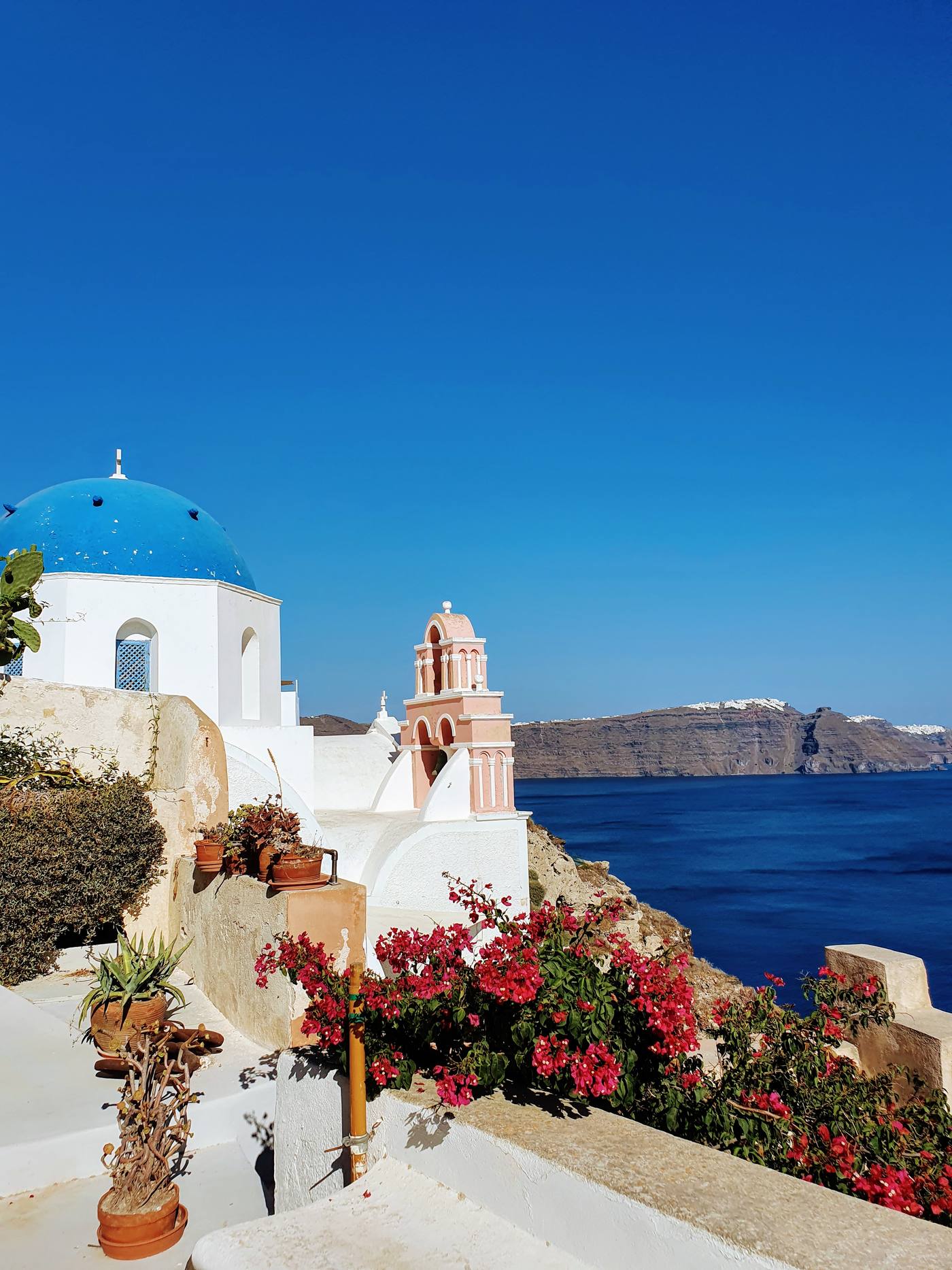 A blue-domed church and pink bell tower overlooking the Santorini caldera, with the volcanic cliffs and white clifftop villages visible across the water