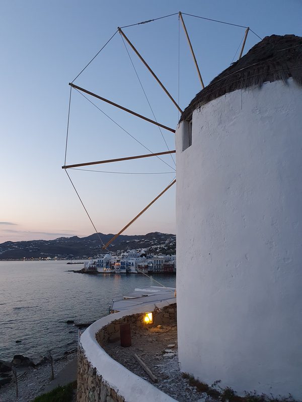Two whitewashed Kato Mili windmills lit at dusk above Mykonos harbour, with Little Venice glowing below