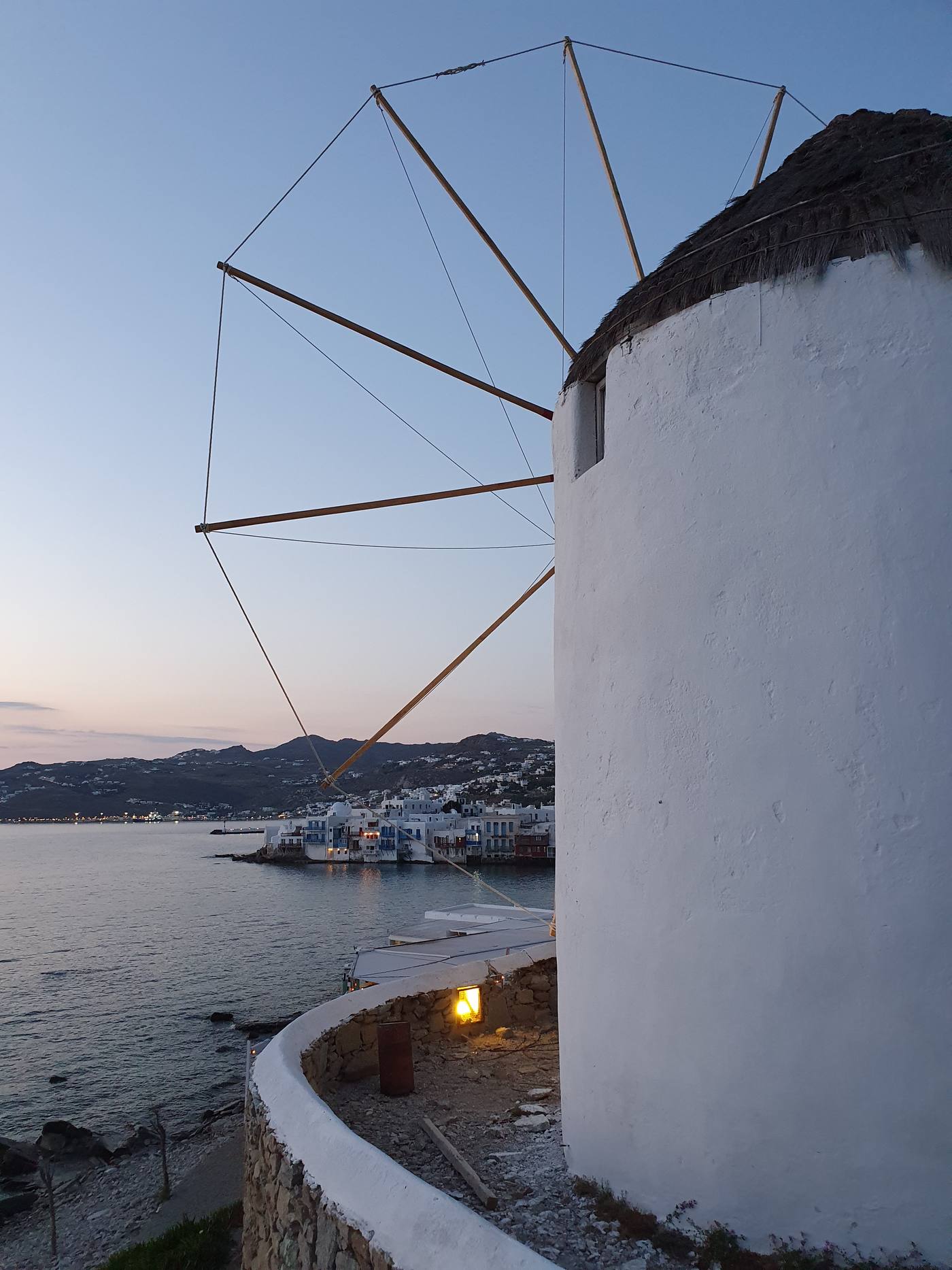 Two whitewashed Kato Mili windmills lit at dusk above Mykonos harbour, with Little Venice glowing below