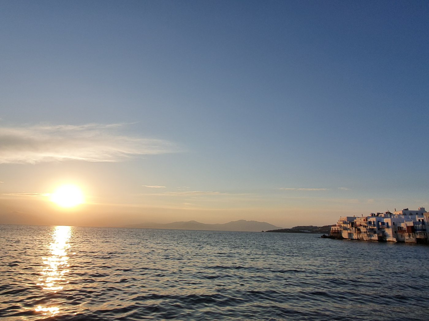 The sun setting over the sea from the Mykonos town waterfront, with the Little Venice buildings just visible on the point